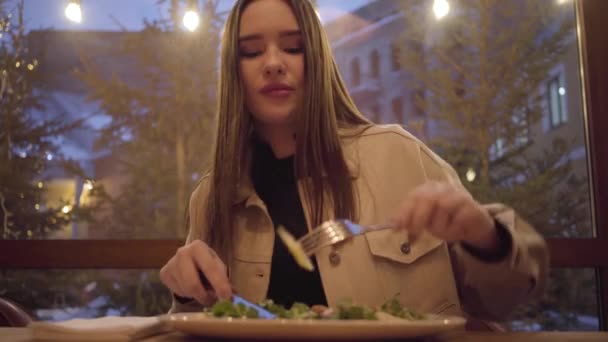 Hezká mladá dívka jíst salát nožem a vidličkou zblízka. Osamělá žena tráví čas mimo domov v restauraci či kavárně — Stock video