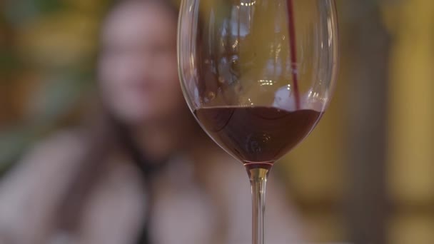 Vino tinto vertiendo lentamente en la copa en primer plano de cerca. Figura borrosa de mujer joven está en el fondo. Movimiento lento . — Vídeos de Stock