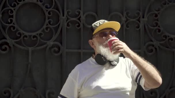 Retrato de un viejo abuelo barbudo vestido a la moda bebiendo café y torciendo su barba gris al aire libre . — Vídeos de Stock