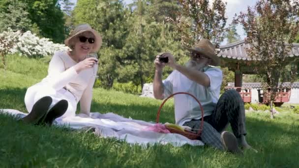 Senior man fotograferen zijn elegantie vrouw op de camera op de picknick in het park — Stockvideo