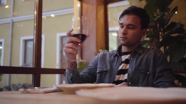 Retrato de um tipo bonito a beber vinho sentado numa mesa num restaurante. Jovem desfrutando de uma bebida enquanto espera por um delicioso jantar . — Vídeo de Stock