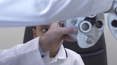 Küçük çocuklar göz muayenesinde, optometrist göz doktoruna phoropter kullanarak kapatın. Doktor beyaz tıbbi ceket elinde teen melez çocuğun gözleri phoropter koyar. Ağır çekim.