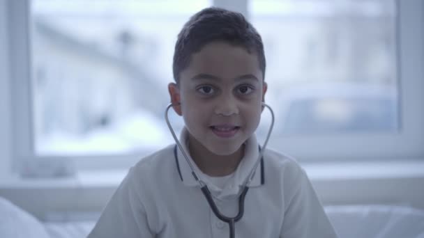 Sztetoszkóp, a fülében a kamera mosolyogva nézett aranyos kisfiú portréja. Egészségügyi ellátás, az egészséges életmód és a koncepció orvosi ügyelet — Stock videók