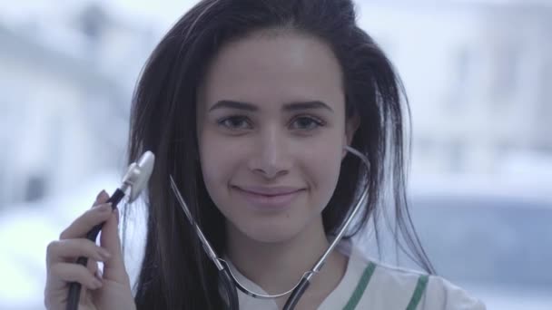 Portrét krásné usměvavé dívky zblízka. Mladá bruneta žena s dlouhými vlasy v fotoaparát drží stetoskop. — Stock video
