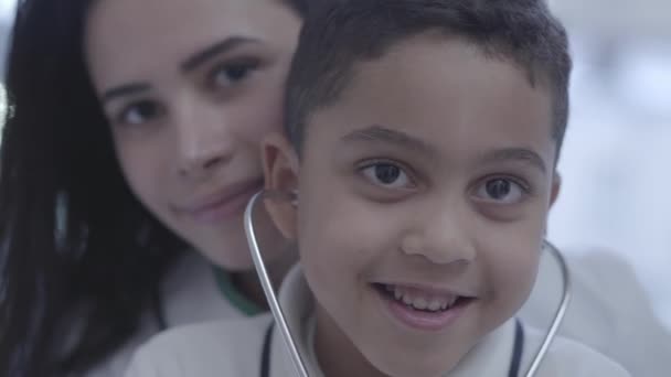 Retrato de una hermosa mujer caucásica joven con un guapo chico mulato mirando en la cámara sonriendo. El niño tiene estetoscopio en las orejas. Concepto de familia multicultural linda feliz. Asistencia sanitaria — Vídeos de Stock