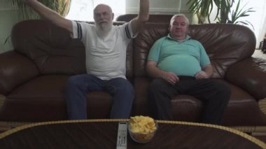 Kahverengi deri kanepe izlerken Tv. sakallı duygusal adam üzerinde oturan iki olgun erkekler futbol takımı fanatiği. Uzaktan kumanda ve cips masaya vardır. İki eski arkadaş evde eğlence