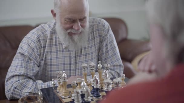 Δύο ηλικιωμένοι, παίζοντας σκάκι στο σπίτι κάθεται στον καναπέ δέρματος. Γενειοφόρος άντρας σκέφτεται ποιο κομμάτι σκάκι για να κάνει μια κίνηση. Καυκάσιος γέροι γείτονες φίλοι παίζουν σκάκι χαρωπά σε εσωτερικούς χώρους — Αρχείο Βίντεο