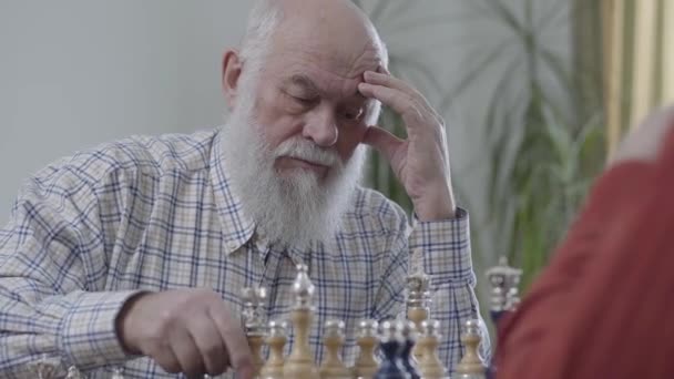 Velhos amigos jogando xadrez sentado em casa no sofá de couro. Homem barbudo pensando que peça de xadrez para fazer um movimento . — Vídeo de Stock