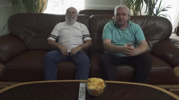 Zwei ältere Männer sitzen auf dem braunen Ledersofa vor dem Fernseher. Fernbedienung und Chips liegen auf dem Tisch. Freizeit zweier alter Freunde zu Hause — Stockvideo