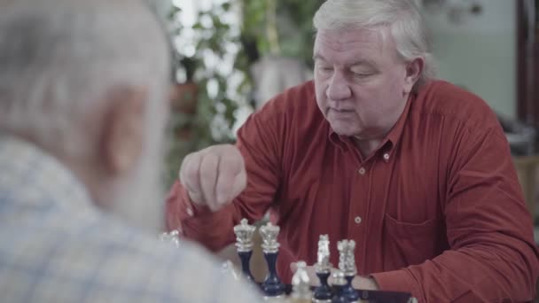 Dos hombres mayores jugando al ajedrez sentados en casa. Un hombre regordete con camisa roja haciendo un movimiento. Caucásico ancianos vecinos amigos jugando ajedrez alegremente en el interior. Concepto de desafío de inteligencia — Vídeo de stock