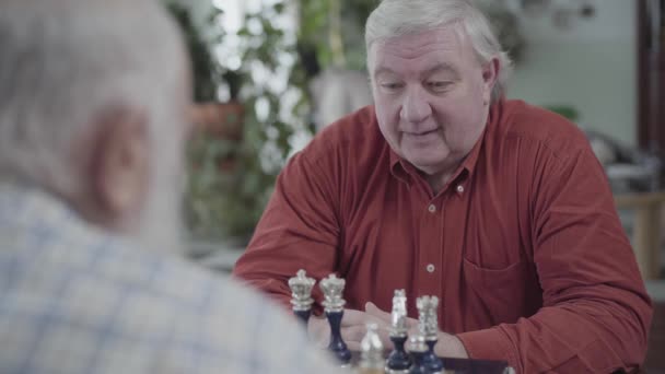 Двое взрослых мужчин играют в шахматы, сидя дома. Кавказские старики соседи радостно играют в шахматы в помещении. Концепция интеллектуального вызова — стоковое видео
