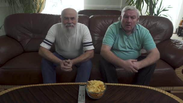 Zwei ältere Männer sitzen auf dem braunen Ledersofa vor dem Fernseher. Freunde lehnen sich gleichzeitig nach vorne und warten auf das Tor, aber das passiert nicht. Fernbedienung und Chips liegen auf dem Tisch — Stockvideo