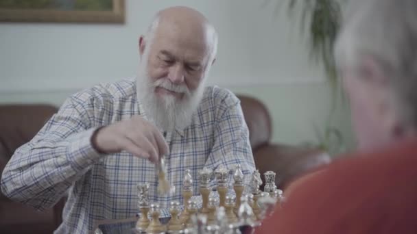 Взрослые друзья играют в шахматы, сидя дома на кожаном диване. Бородатый человек думает, какую шахматную фигуру сделать ход . — стоковое видео