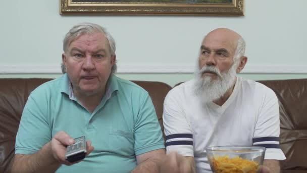 Portrait of two mature senior men sitting on the brown leather sofa watching TV. One man holding the bowl with chips, other witches channels using the remote. Leisure of the pensioners — Stock Video