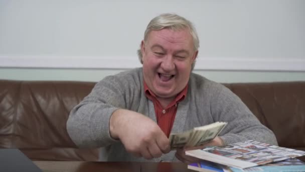 Szczęśliwy Starzec wyrzucać dolarów siedział przy stole drewniane z bliska. Pozytywny człowiek bogaty pokazuje swoje pieniądze. Zabawny dziadka na relaks w domu siedzi na skórzanej kanapie. Wypoczynek — Wideo stockowe