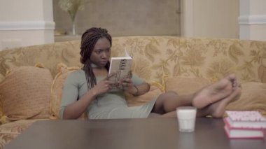 Koç üzerinde oturma odasında oturma ve ilginç bir kitap okuma güzel Amerikan Afrika kadın