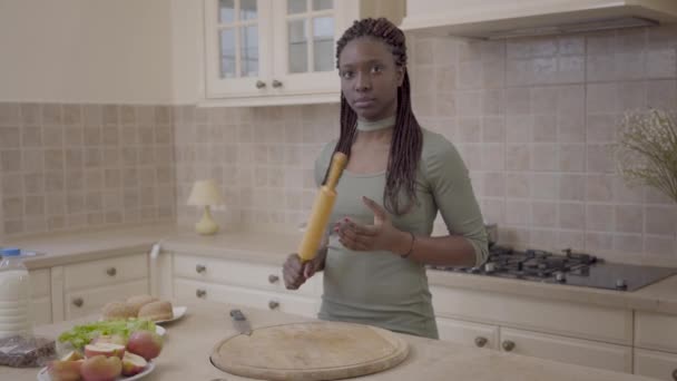 Портрет довольно африканской американки с дредами, держащими катушку из теста, стоящую на кухне и смотрящую в камеру. Камера движется влево — стоковое видео