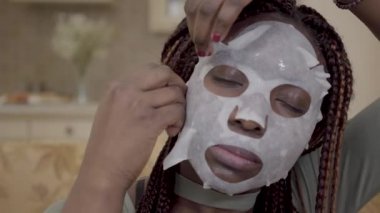 Gençleştirme yüz maskesi yüzünde uygulayarak genç Afro-Amerikan kadın portresi evde kapatın. Bayan güzellik ve cilt sağlığı hakkında umurunda değil.