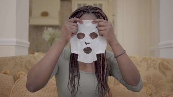 Retrato de mulher americana africana bonita colocando máscara facial hidratante em seu rosto no treinador na sala de estar moderna — Vídeo de Stock