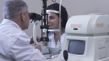 Güzel genç kadın göz test makinesi bakarak ve gülümseyen beyaz tıbbi ceket Doktor önünde oturan klinikte göz testi yapmak. Ofiste hasta muayene olgun optometrist. Kamera sağa hareket ediyor