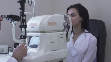 Oldukça genç kadın, modern göz testi makinesinde görünümlü beyaz tıbbi ceket Doktor önünde oturan klinikte göz testi yapmak. Ofiste hasta muayene olgun optometrist
