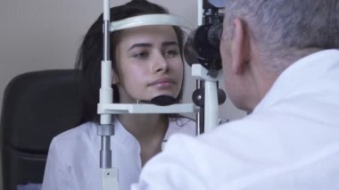 Güzel genç kadın göz test makinesi bakıyor ve gülümseyen beyaz tıbbi ceket Doktor önünde oturan klinikte göz testi yapmak. Ofiste hasta muayene olgun optometrist. Kamera sağa hareket ediyor