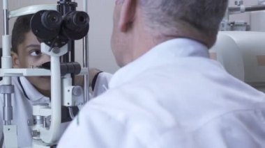 Klinikte küçük yakışıklı çocuk Doe göz testi göz test makinesine bakarak beyaz tıbbi ceket Doktor önünde oturuyor. Olgun optometrist ışık kullanarak hasta gözü inceliyor. Kamera sola hareket ediyor