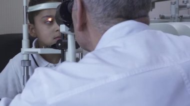 Klinikte küçük yakışıklı çocuk Doe göz testi göz test makinesine bakarak beyaz tıbbi ceket Doktor önünde oturuyor. Olgun optometrist ışık kullanarak hasta gözü inceliyor. Kamera sola hareket ediyor