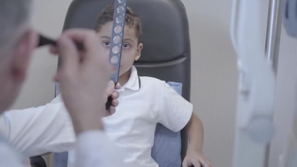 Porträt eines gutaussehenden Afroamerikaners, der auf dem Stuhl des Augenarztes sitzt. Ein professioneller Augenarzt überprüft die Sehkraft des Kindes. Augenuntersuchung. Kamera rückt hinter Arzt zurück — Stockvideo