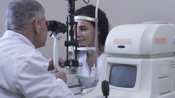 Mulher bonita fazer teste de olho na clínica sentado na frente do médico em casaco médico branco olhando na máquina de teste de olho e sorrindo. Otometrista maduro examinando paciente no consultório. Câmara se move para a direita — Vídeo de Stock