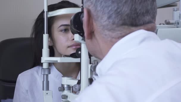 Ganska ung kvinna gör ögontest på kliniken sitter framför läkare i vit medicinsk päls söker i ögat test maskin och leende. Mogna optiker undersöka patienten i ämbetet. Kameran rör sig rätt — Stockvideo