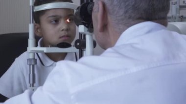 Klinikte küçük yakışıklı çocuk Doe göz testi göz test makinesine bakarak beyaz tıbbi ceket Doktor önünde oturuyor. Olgun optometrist ışık kullanarak hasta gözü inceliyor. Kamera sola hareket ediyor