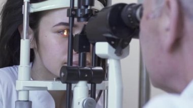 Göz testi makinesi bakarak beyaz tıbbi ceket Doktor önünde oturan klinikte göz testi yapan güzel genç kadın. Olgun optometrist ışık kullanarak hasta gözü inceliyor. Sağlık