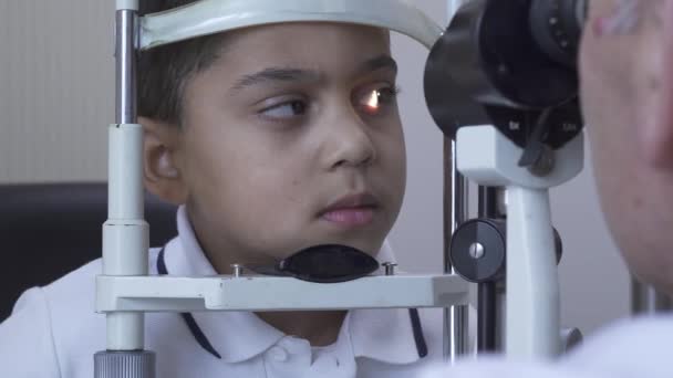 Malý pohledný kluk na klinice sedí před doktorem v bílém lékařském kabátě a dívá se do očí. Zralý optik zkoumáním pacientova oka pomocí světla. Kamera se pohybuje doleva — Stock video
