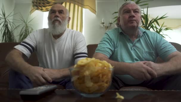 Két érett vezető férfi ül a barna bőr kanapé tévénézés. a nyugdíjasok pihenése — Stock videók