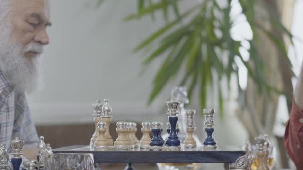 Deux amis aînés matures jouant aux échecs assis à la maison. Vieux voisins caucasiens jouant aux échecs joyeusement à l'intérieur. Concept de défi du renseignement — Video