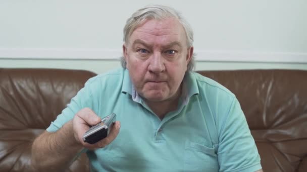 Porträt eines erwachsenen Mannes in blauem T-Shirt, der auf dem braunen Ledersofa vor dem Fernseher sitzt. alter Mann schaltet Kanäle mit der Fernbedienung. Freizeit des Rentners — Stockvideo