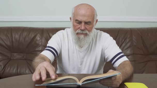 Hombre adulto barbudo reflexivo sentado en la mesa de madera en el sofá de cuero leyendo el libro. Concepto educativo — Vídeo de stock