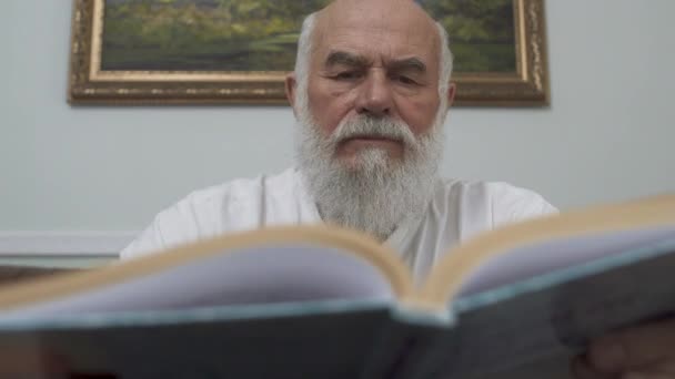 Close-up portret nadenkende bebaarde Senior man zittend aan de houten tafel op lederen sofa het lezen van het boek. Kaukasische oude man genieten van tijd binnenshuis. Vrije tijd van de gepensioneerde. Onderwijsconcept — Stockvideo