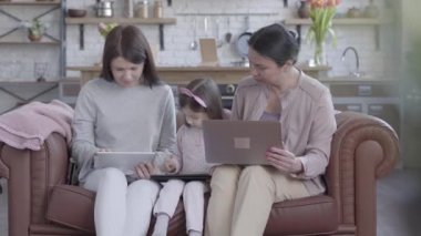 Büyükanne, anne ve küçük kızı modern stüdyo dairede deri kanepede birlikte oturuyor. Herkes Tur, laptop veya tablet üzerinde Gadget var.