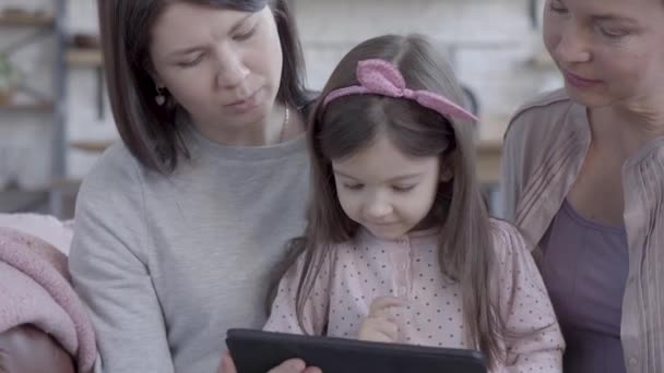 Drei Generationen von Frauen sitzen zusammen auf dem Sofa, unterhalten sich und beobachten, wie kleine Mädchen Spiele mit Tablets spielen. — Stockvideo