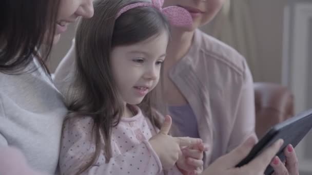 Trois générations de femmes assises ensemble dans le canapé, parlant et regardant comment les petites filles jouent au jeu sur tablette . — Video