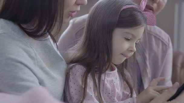 Mormor, mor och liten dotter sitter tillsammans på soffan i modern lägenhet. Flickan håller gadget, mamma och farmor sitter nära och titta på skärmen leende. Begreppet generation. — Stockvideo