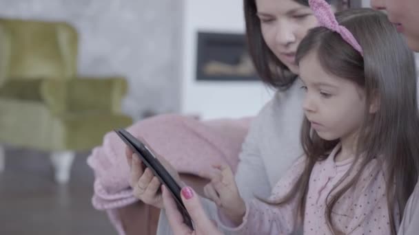 Τρεις γενιές γυναικών κάθονται μαζί στον καναπέ, μιλούν και να εξηγήσει σε ένα μικρό αστείο κορίτσι πώς να παίξει το παιχνίδι στο tablet. — Αρχείο Βίντεο