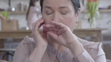 Ön planda deri kanepede otururken elleri ile kafa tutan renkli siyah saçlı kadın yorgun hasta kıdemli portresi. Anne ve kızı arka plan bulanık rakamlar.