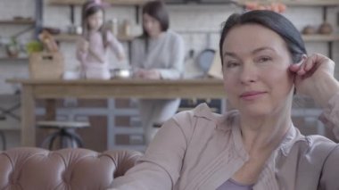 Kamera ön planda deri kanepede oturan renkli siyah saç seyir ile mutlu kendine güvenen kadın kıdemli portresi. Anne ve kızı arka plan bulanık rakamlar.