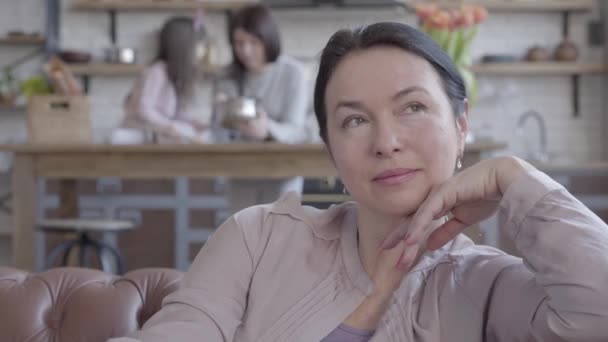 Retrato mulher sênior sentado no sofá, lembre-se de dias passados com sorriso e obrigado. No fundo é sua filha ensinando como cozinhar sua pequena menina na cozinha — Vídeo de Stock