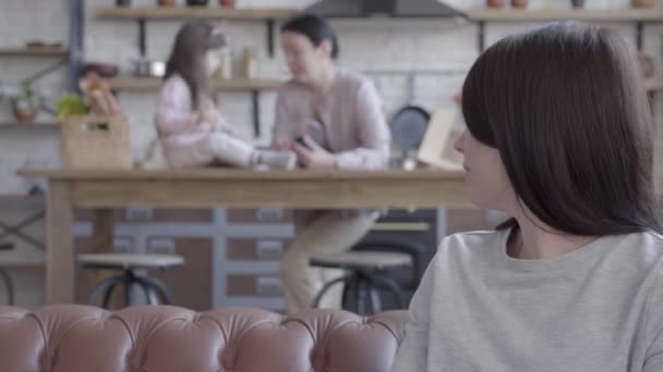 Retrato de mujer feliz confiada mirando hacia atrás a la pequeña hija jugando con la abuela y luego girando a la cámara. La señora está sentada en el sofá de cuero en primer plano. Concepto de generación — Vídeo de stock