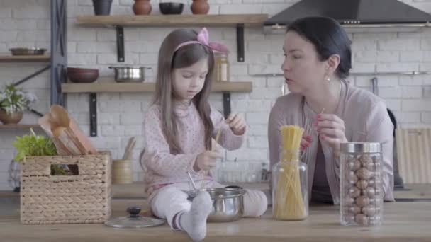 Pequeña chica divertida sentada en la mesa ayudando a su abuela a cocinar fideos. Mujer mayor de pie cerca de la mesa con tarros de nueces y fideos en la superficie. Concepto de generación, amor — Vídeos de Stock