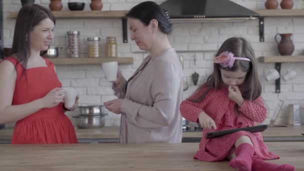 Tre generazioni di donne in studio che bevono te 'insieme e parlano. Incredibile piccola ragazza giochi in tablet sul tavolo della cucina . — Video Stock